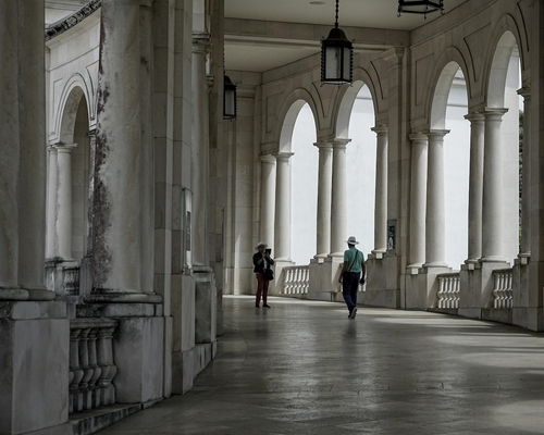 Überdachter Säulengang mit Rundbögen im Heiligtum von Fátima, zwei Personen gehen in Richtung des Ausgangs, Lampen hängen von der Decke.