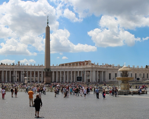 Weitwinkelansicht des Petersplatzes mit Menschen, dem zentralen Obelisken, Brunnen und der Fassade des Petersdoms unter blauem Himmel mit Wolken.