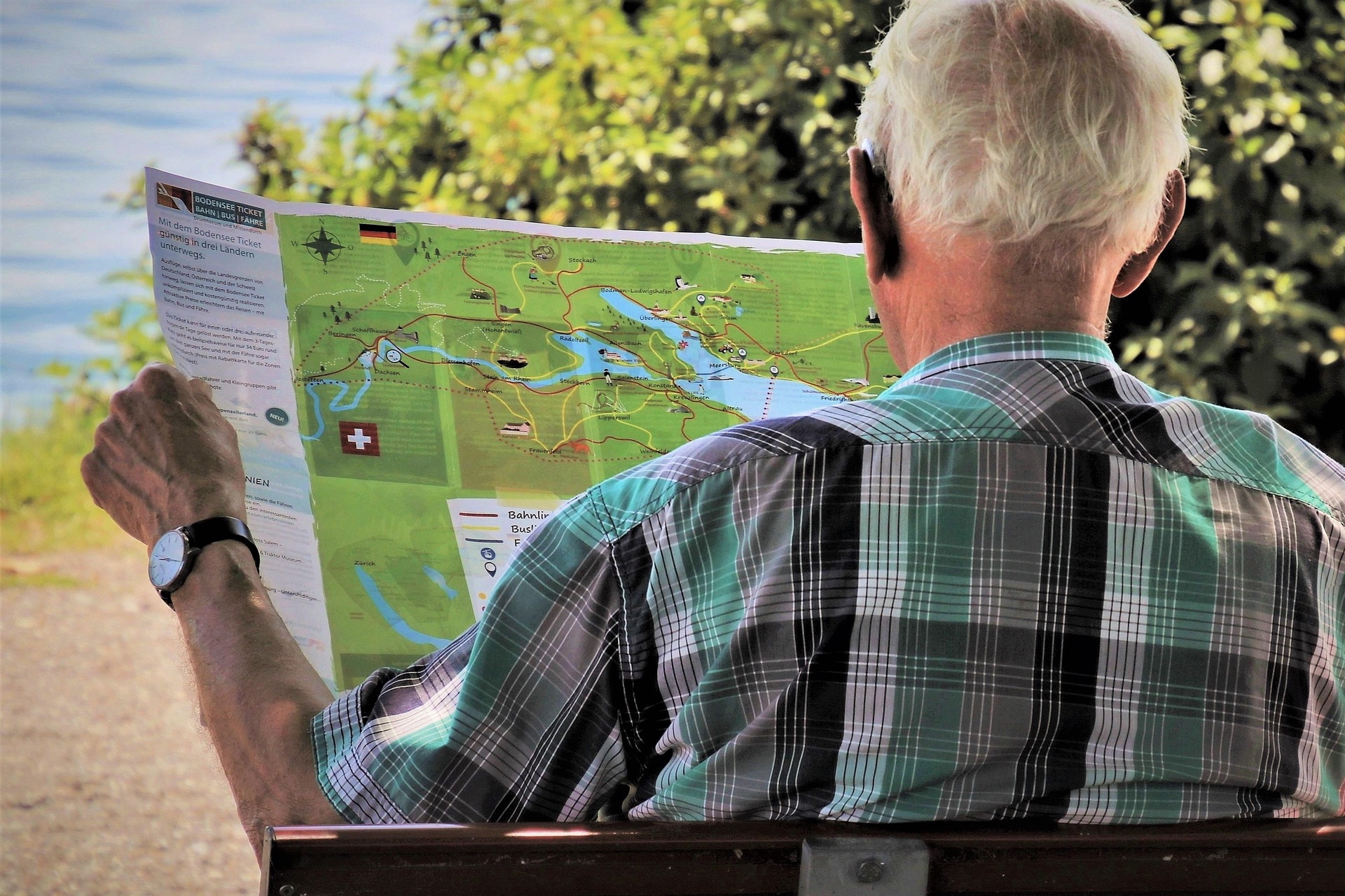 Älterer Mann mit kariertem Hemd sitzt auf einer Bank und betrachtet eine großformatige Landkarte mit Grenzverlauf zwischen Deutschland und der Schweiz, im Hintergrund Natur und Wasser.